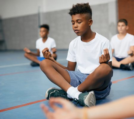 African American schoolboy and his friends meditating in lotus position during PE class at school gym.